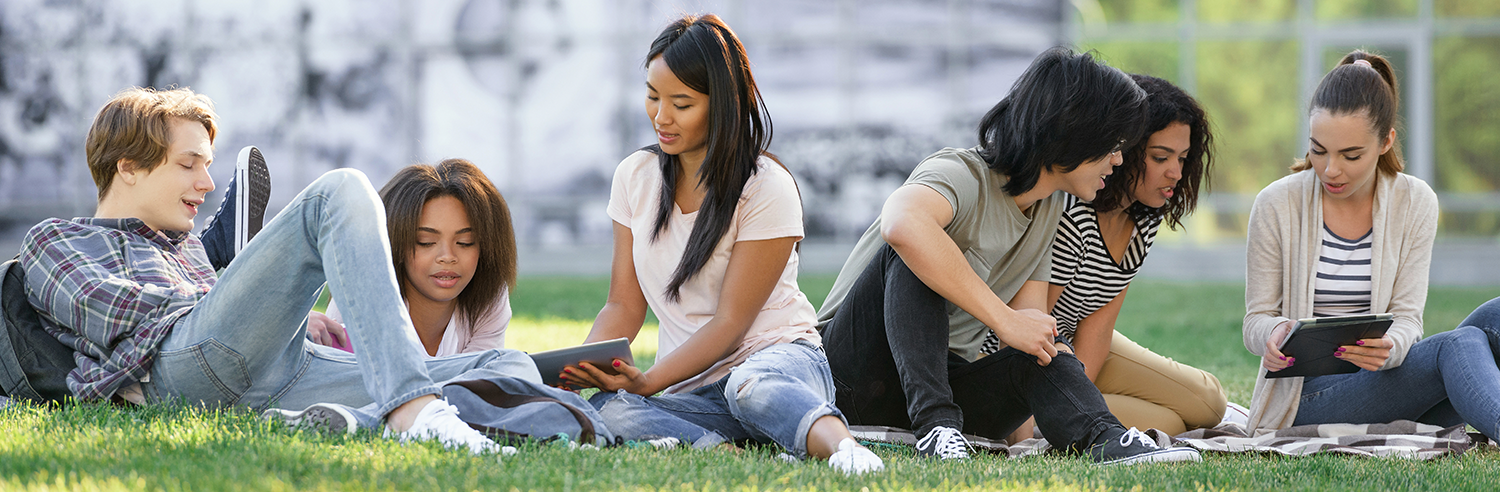 A group of young women and men students sitting on a lawn talking with each other and looking at computers.